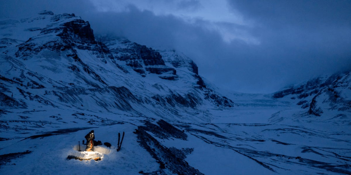 A solo backcountry skier sets up camp in a snowy, mountainous area at twilight