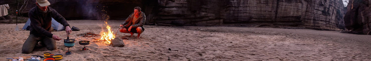 two men sit around a fire, cooking dinner at their campsite