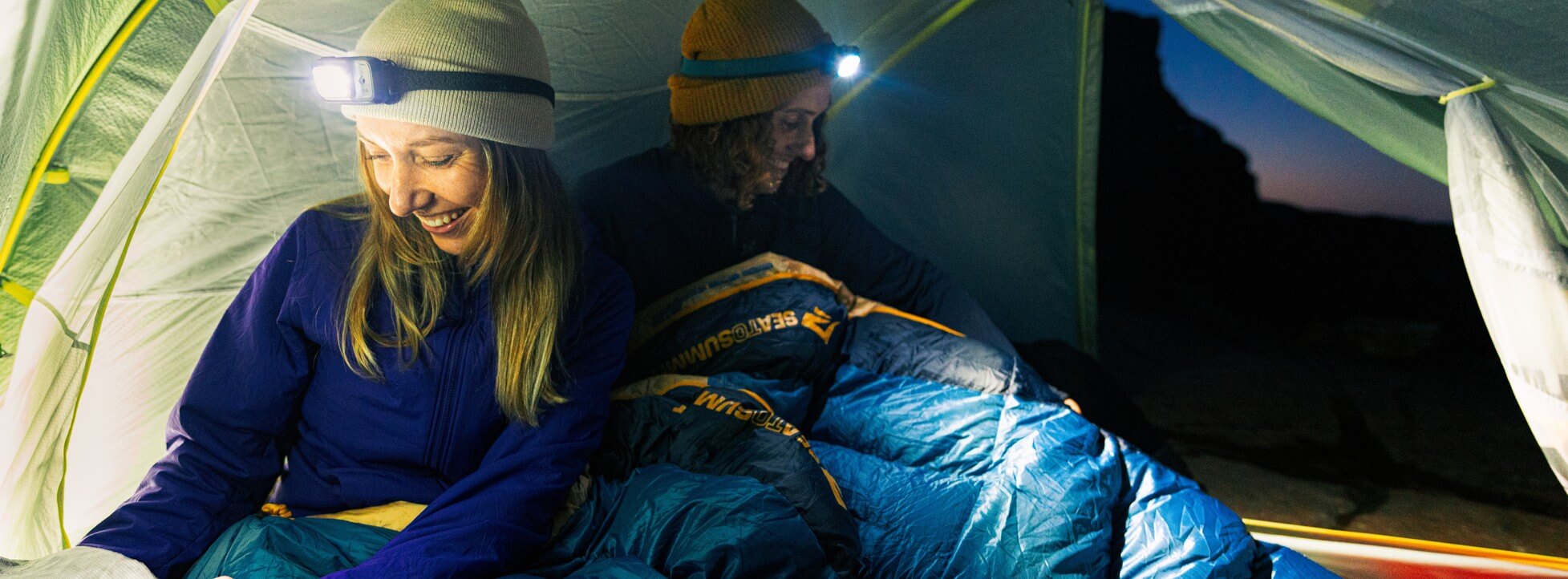 Two campers wearing headlamps sit in blue sleeping bags inside a tent at dusk.
